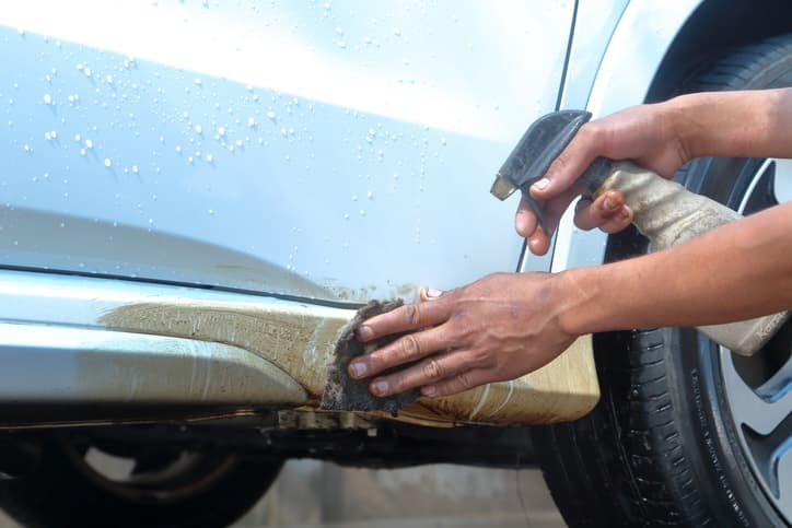 Close-up of hands cleaning paint off a car with a sponge and spray bottle, focusing on the lower section of the vehicle near the wheel.