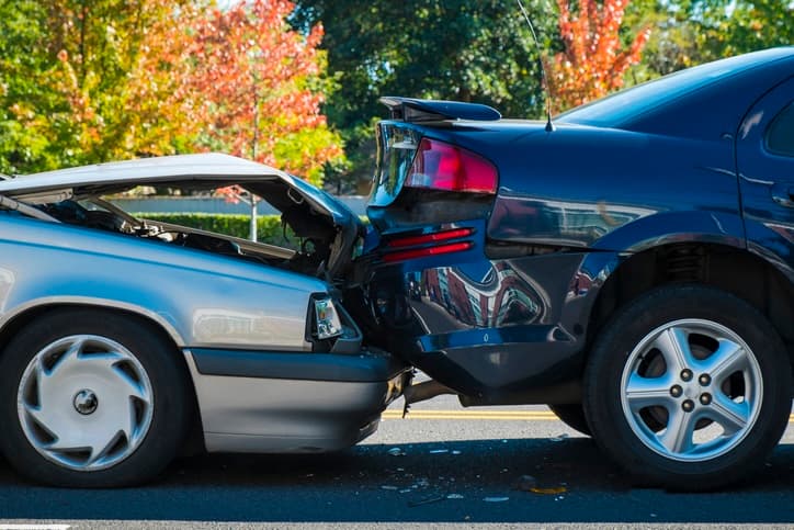 A rear-end collision between two cars on a sunny day, showing significant damage to the front of a silver car and the rear of a dark blue car, with autumn trees in the background.